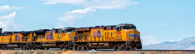 Salt Lake City, USA - July 27, 2019: Utah landscape with train on railroad with cars containers, and american flag on yellow color locomotive with mountains in desert background