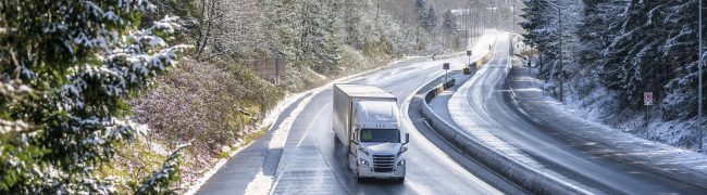 White modern bonnet popular professional big rig semi truck with dry van semi trailer going on the wet dangerous slippery icy winter road with snow on the trees on the sides of the divided highway