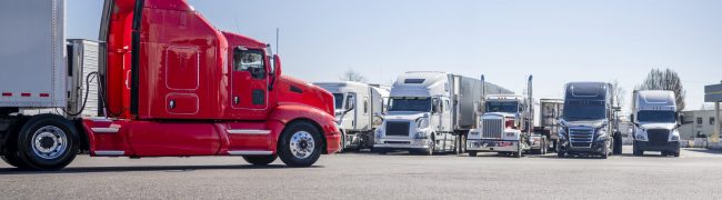 Long haul bright red big rig semi truck with extended cab transporting cargo in dry van semi trailer driving on truck stop parking lot with direction of movement arrow and row of another semi trucks