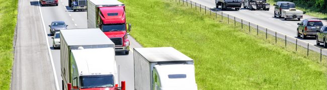 A heavy flow of 18-wheelers peppered with cars and SUVs roll down a Tennessee interstate highway.  Heat rising from the pavement gives a shimmering effect to vehicles and background in the distance.