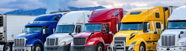 Different make big rigs semi trucks tractors with loaded semi trailers standing in the row on truck stop parking lot at early morning waiting for the route continuation time according to the log book